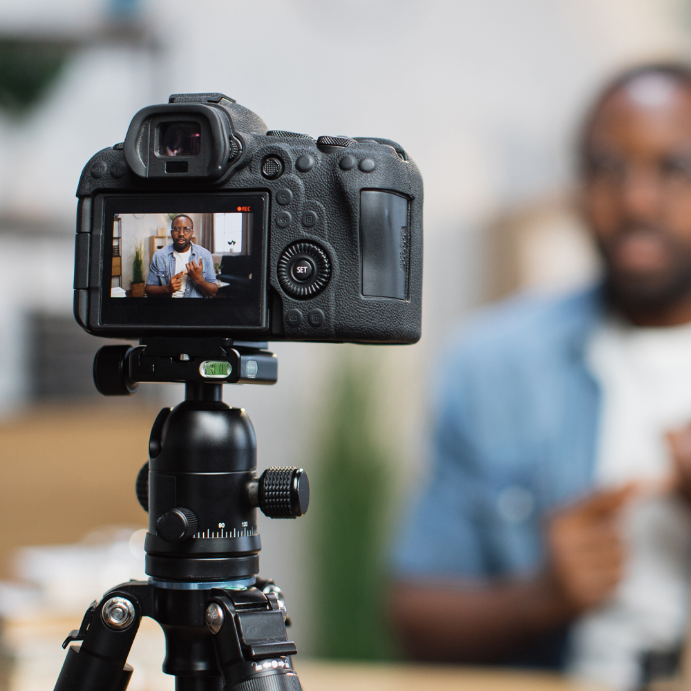 Blur background of african guy in casual wear recording new video blog while sitting at home workplace. Focus on digital camera fixed in tripod. Concept of people and social networks.